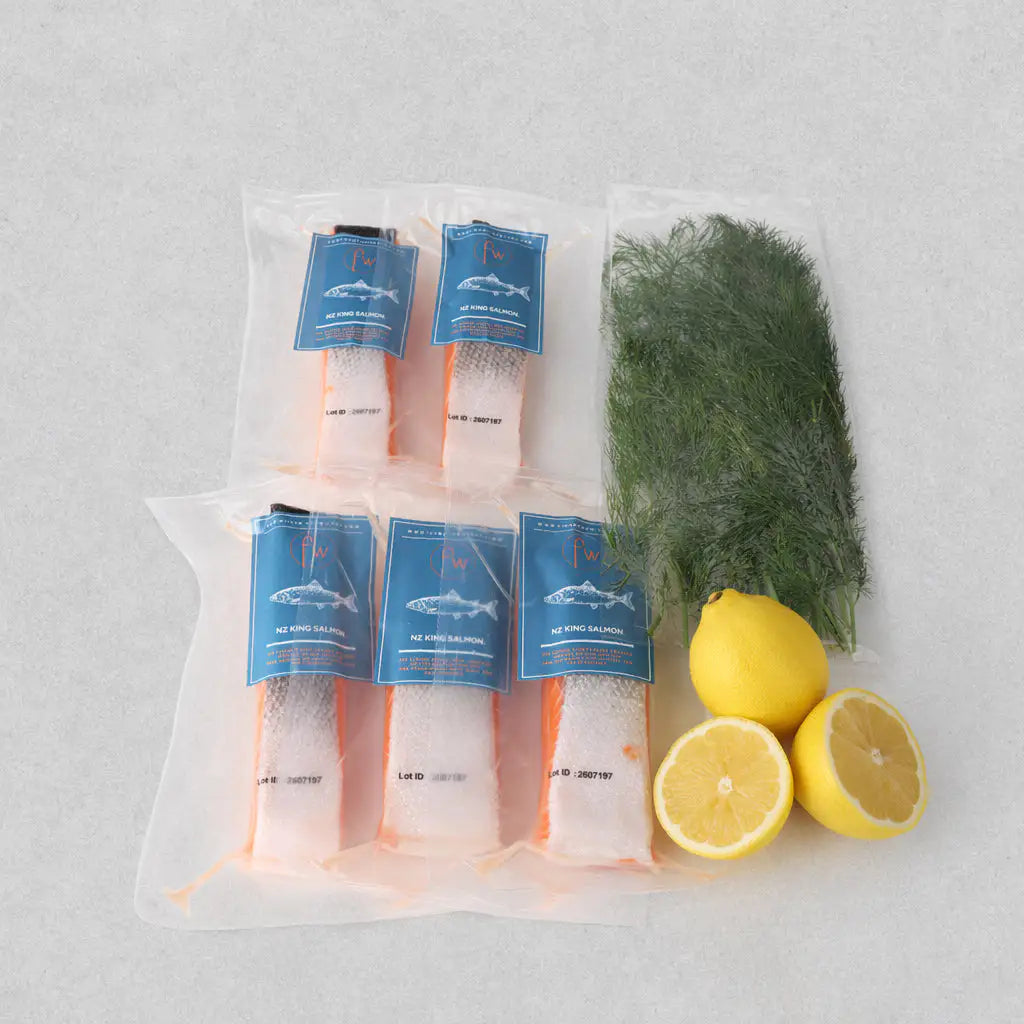 Six vacuum-sealed packages of salmon fillets with blue labels, arranged on a light gray surface alongside a bag of dill and lemon garnish.