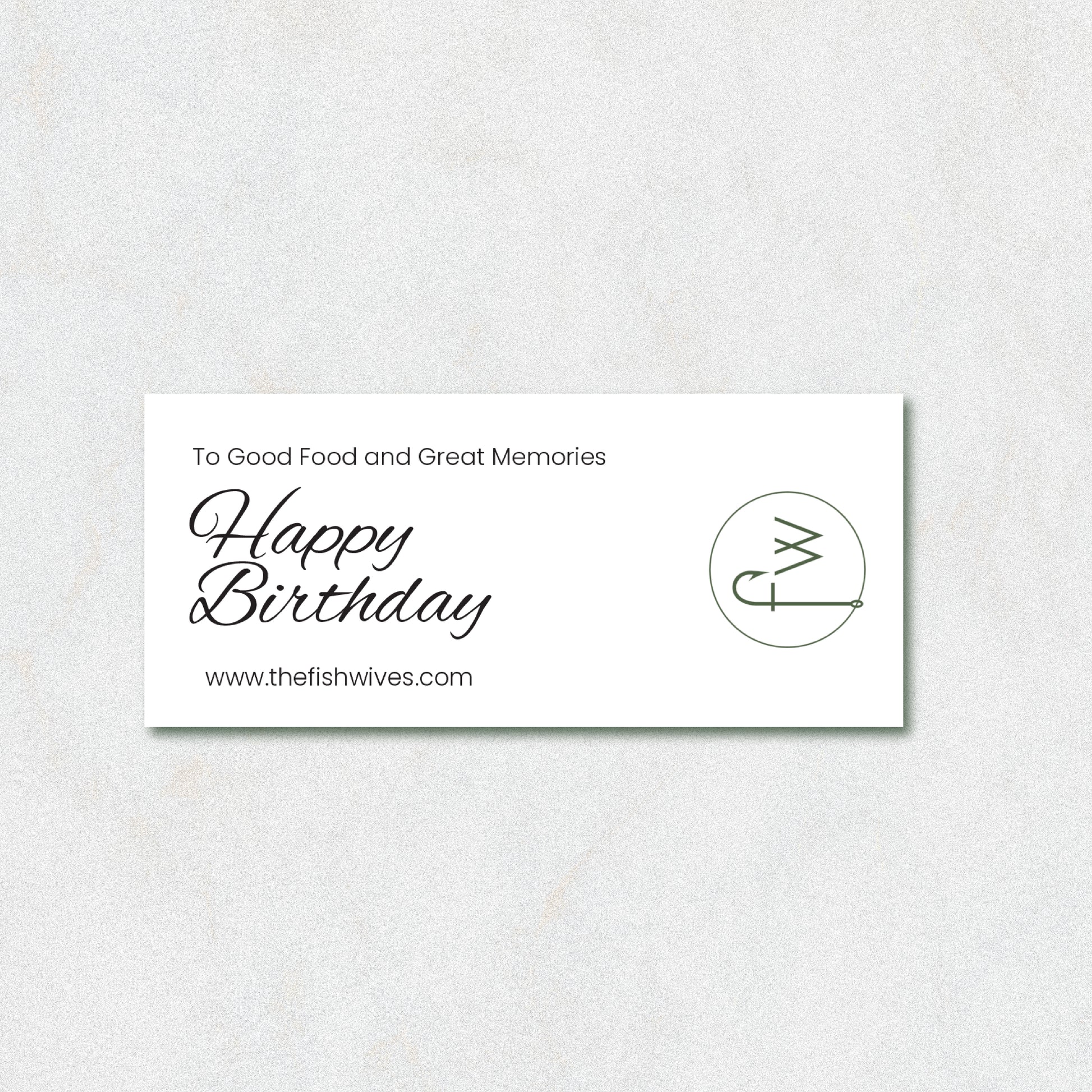 A rectangular white card with black text reading 'To Good Food and Great Memories Happy Birthday' and 'www.thefishwives.