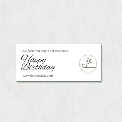 A rectangular white card with black text reading 'To Good Food and Great Memories Happy Birthday' and 'www.thefishwives.