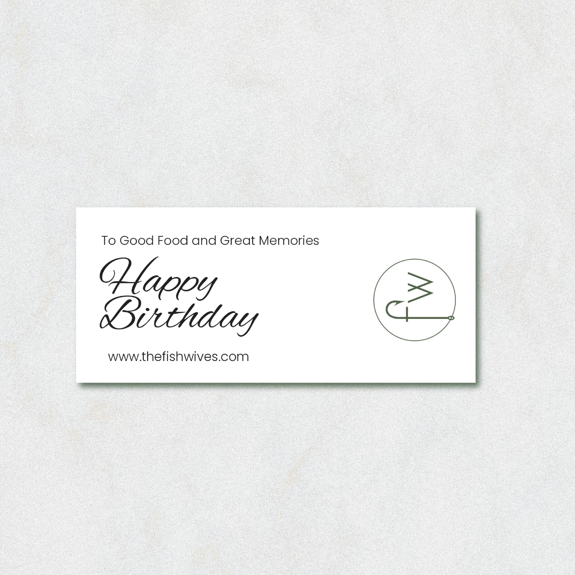 A rectangular white card with black text reading 'To Good Food and Great Memories Happy Birthday' and 'www.thefishwives.