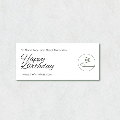 A rectangular white card with black text reading 'To Good Food and Great Memories Happy Birthday' and 'www.thefishwives.