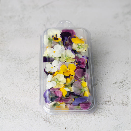 A clear plastic clamshell container filled with colorful edible flower petals, including yellow, purple, and white pansies, set on a light gray textured surface.
