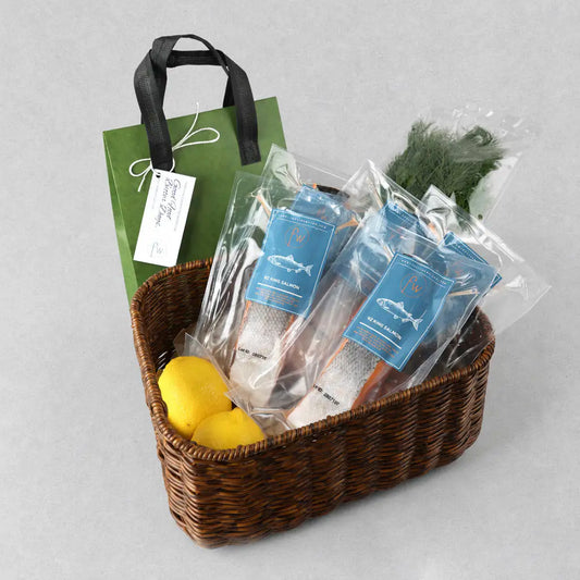 A gift basket set on a light gray background, containing a brown woven basket with two packaged salmon fillets, two yellow lemons, a green gift bag with a white tag, and a small bunch of dill.
