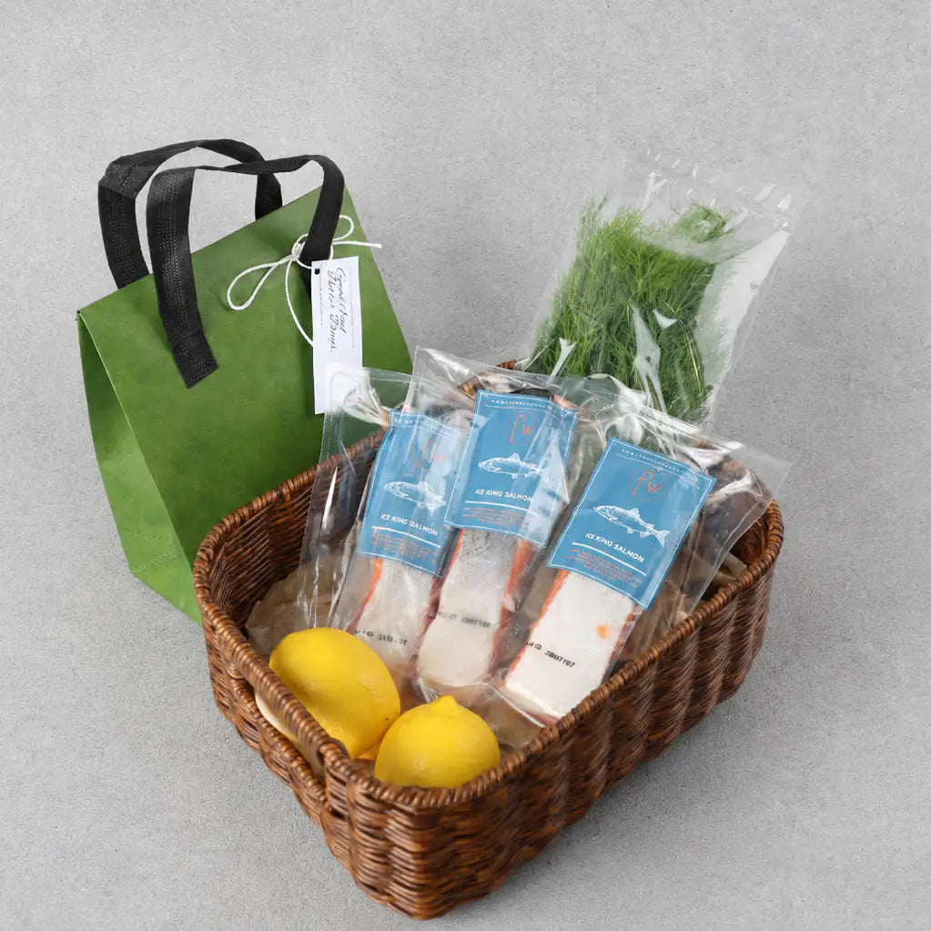 A gift basket set on a gray background, featuring a brown woven basket with lemons, packaged salmon fillets with blue labels, a bag of dill, and a green paper gift bag with black handles and a white tag.