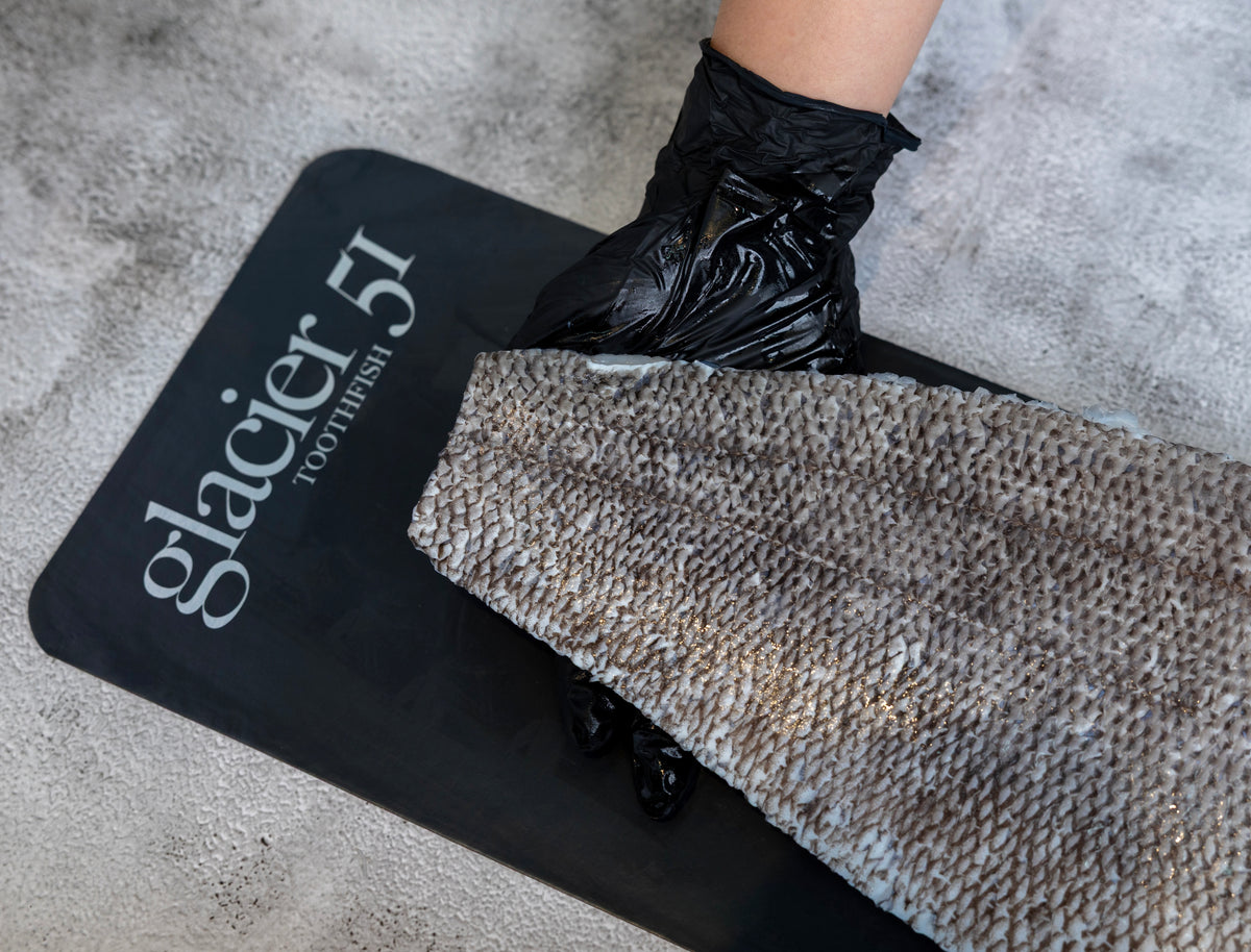 A close-up of a textured gray and white toothbrush being wiped across a black Glacier 51 toothbrush mat by a hand wearing a black plastic glove, set on a gray concrete floor.