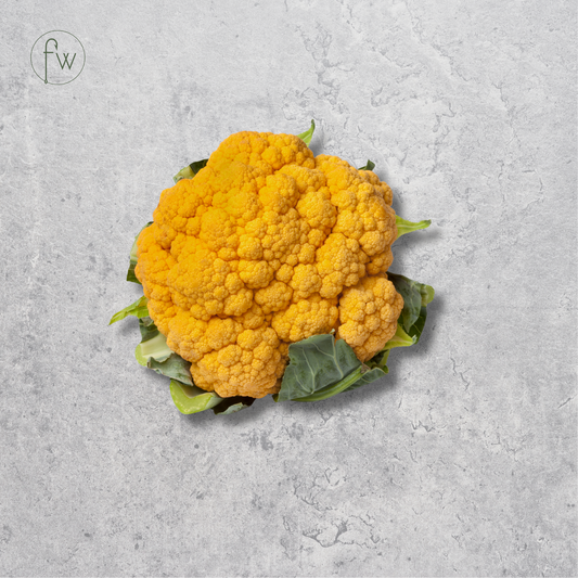 A single bright yellow-orange cauliflower head with green leaves, photographed from above on a light gray textured surface. A small circular 'fw' logo appears in the upper left corner.