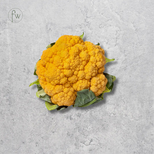 A single bright yellow-orange cauliflower head with green leaves, photographed from above on a light gray textured surface. A small circular 'fw' logo appears in the upper left corner.