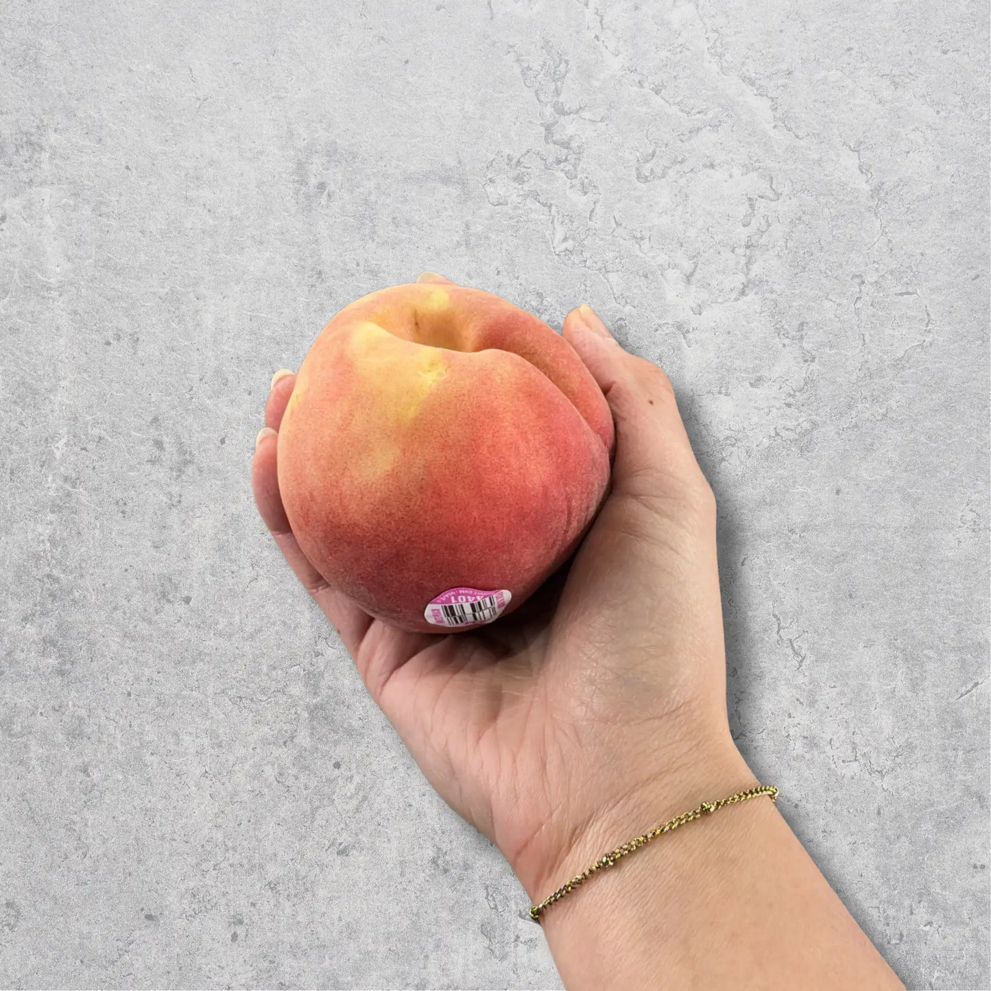 A hand holding a ripe, fuzzy peach with a small sticker on its side, set against a textured gray concrete background.
