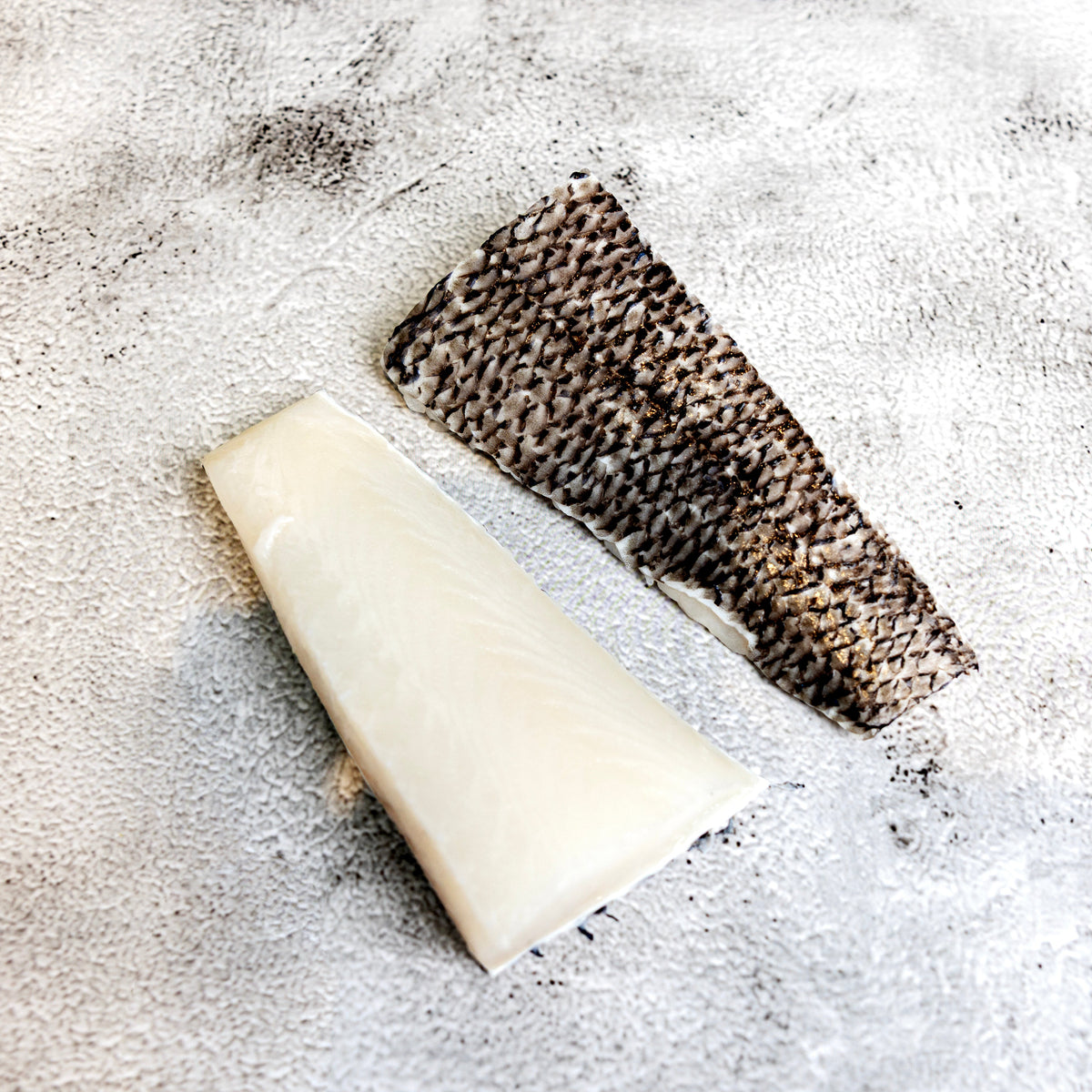 A wedge of pale cream-colored cheese next to a piece of dark brown and black textured fish skin, arranged on a light gray textured surface.